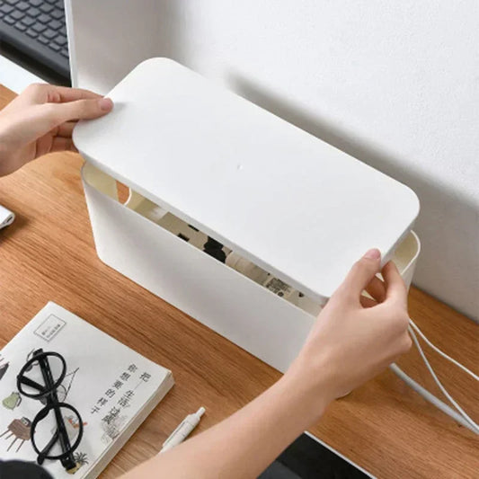 A hand lifting the lid off a white cable management box on a wooden desk, showing the power strip and coiled cable inside.