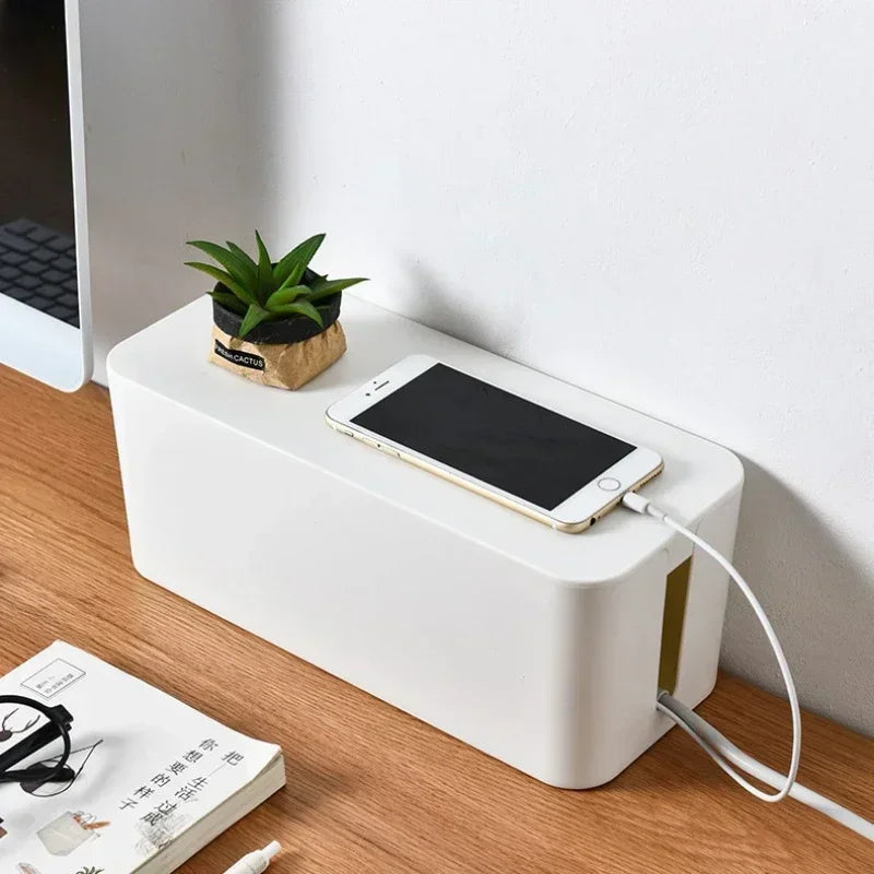A white cable management box on a light oak desk with a phone resting on the lid charging through a side opening, a small potted succulent beside it and a monitor behind.