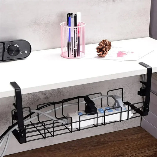 A black wire cable tray fixed under a white desk, holding a power strip and gathered cables, with a pen pot and pine cones on the desk above.