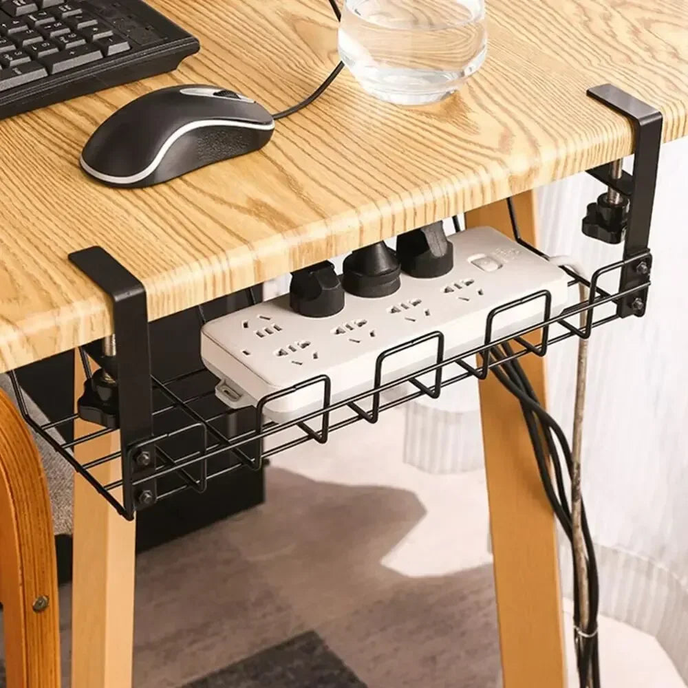 A black wire cable tray clamped beneath a solid oak desk holding a white power strip and its plugs, with a computer mouse and a glass of water on the desk above.