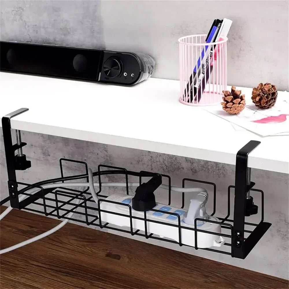 A side view of a black wire cable tray clamped under a white desk with cables inside, a desk speaker and a pen pot visible above.